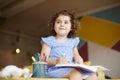 Beautiful baby girl with dark curly hair in blue dress dreamily looking aside while drawing in coloring book with felt