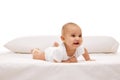 Beautiful baby, little girl in white comfortable clothes lying on belly on bed with pillow behind against white Royalty Free Stock Photo