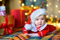 Baby in Santa Claus costume lies on the floor near the presents on the background of bright festive garlands