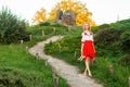 Beautiful barefoot woman in stylish red white dress holding shoes in hand and walking on path at hill. standing on road village