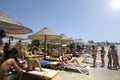 Beautiful beach resort with straw umbrellas on a blue sky and white clouds. On the background some people having fun on the beach