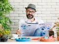 Beautiful beard man wearing hat and eyeglasses sitting in home indoor garden and reading newspaper with wite coffee cup Royalty Free Stock Photo