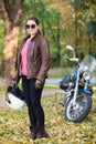 A beautiful biker girl holding helmet next ner chopper bike, autumn park, full length portrait