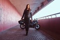 A beautiful biker girl holding helmet next superbike walks on a sidewalk inside the bridge.