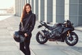 A beautiful biker girl holding helmet next superbike outside a building.