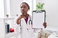 Beautiful black doctor woman holding clipboard serious face thinking about question with hand on chin, thoughtful about confusing