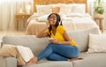 Beautiful black woman with mobile phone and headphones sitting on comfy sofa and listening to music at home