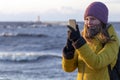 Beautiful blond caucasian girl in winter hat and bright coat looking at her mobile phone at the sunset seaside Royalty Free Stock Photo