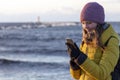 Beautiful blond caucasian girl in winter hat and bright coat looking at her mobile phone at the sunset seaside Royalty Free Stock Photo