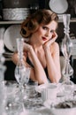 Beautiful blond girl with vintage make up and hairstyle in evening shining dress sitting at table with plates and glasses