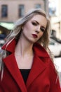A beautiful blond girl wearing a red coat and gloves stands near the barrier among old buildings. Fashionable, industrial, Royalty Free Stock Photo