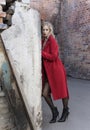 A beautiful blond girl wearing a red coat and gloves stands by a ruined wall and brick arch near an old building. Fashionable, Royalty Free Stock Photo