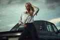 Beautiful blond lady in black striped high waisted pants and oversized white blouse standing at her old car looking away