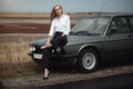 Beautiful blond lady in black striped high waisted pants, white blouse and high heeled shoes sitting on the hood of her old car Royalty Free Stock Photo