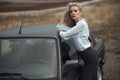 Beautiful blond lady in black striped high waisted pants and white blouse leaning on the top of her old car