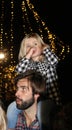 Beautiful blond little girl with her thumb in her mouth and a plaid shirt riding on her fathers shoulders with Christmas lights be