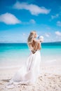 Beautiful blond long hair fiancee in a long white open back wedding dress stand on the white sand beach with a pearl. Looking to Royalty Free Stock Photo