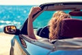 Beautiful blond smiling young woman in convertible top automobile looking sideways while parked near ocean waterfront Royalty Free Stock Photo