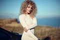Beautiful blond woman with afro curly hair wearing white fitted dress leaning on her automobile with self-confident look