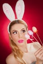 Beautiful blond woman as Easter bunny with rabbit ears on red background, studio shot. Young lady holding three colored eggs Royalty Free Stock Photo