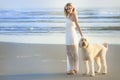 A beautiful blond woman on the beach in a white dress with her Goldendoodle dog.