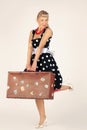 Beautiful blond woman in pinup style dressed in a polka-dot dress is standing and flirtatiously holding a brown suitcase, white Royalty Free Stock Photo