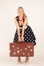 Beautiful blond woman in pinup style, dressed in a polka-dot dress, stands and hardly holds a heavy suitcase, white background Royalty Free Stock Photo