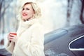 Beautiful blond woman wearing luxurious white fur coat drinking hot coffee on snowy winter day and laughing