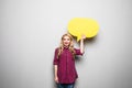 Beautiful blond young woman holding yellow blank speech bubble over grey background Royalty Free Stock Photo