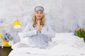 Beautiful blond and young woman, wearing sleeping mask, sitting in bed and doing yoga exercises in the morning Royalty Free Stock Photo