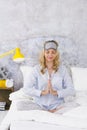Beautiful blond and young woman, wearing sleeping mask, sitting in bed and doing yoga exercises in the morning Royalty Free Stock Photo