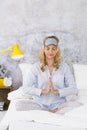 Beautiful blond and young woman, wearing sleeping mask, sitting in bed and doing yoga exercises in the morning Royalty Free Stock Photo