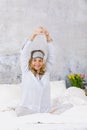 Beautiful blond and young woman, wearing sleeping mask, sitting in bed and doing yoga exercises in the morning Royalty Free Stock Photo