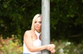 Beautiful blonde adult woman holding and leaning against a lamppost in the park. The woman is sad and looks in pain, anguish and