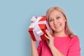 A beautiful blonde adult woman shakes a gift box next to her ear trying to guess what`s inside standing on a blue background