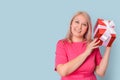 A beautiful blonde adult woman shakes a gift box next to her ear trying to guess what`s inside standing on a blue background, cop
