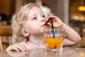 A beautiful blonde baby girl is sitting at a table in a cafe and drinking juice through a straw. Royalty Free Stock Photo