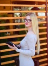 A beautiful blonde in a beige dress and wearing glasses is using a tablet near a wooden wall. Model girl looks at the camera.