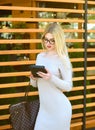 A beautiful blonde in a beige dress and wearing glasses is using a tablet near a wooden wall. Online shopping.