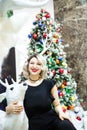 Beautiful blonde in a black dress hugs the figure of a deer near the Christmas tree Royalty Free Stock Photo