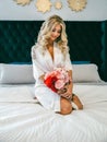 Beautiful blonde with a bouquet of flowers sits on a bed in a white room Royalty Free Stock Photo
