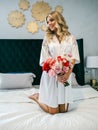 Beautiful blonde with a bouquet of flowers sits on a bed in a white room Royalty Free Stock Photo