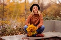 Beautiful blonde in a brown warm sweater, black felt hat, blue jeans and boots sits on a bench in the autumn in the park, holds a Royalty Free Stock Photo