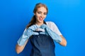 Beautiful blonde caucasian woman wearing cleaner apron and gloves smiling in love doing heart symbol shape with hands
