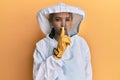 Beautiful blonde caucasian woman wearing protective beekeeper uniform asking to be quiet with finger on lips