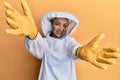 Beautiful blonde caucasian woman wearing protective beekeeper uniform looking at the camera smiling with open arms for hug