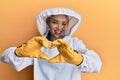 Beautiful blonde caucasian woman wearing protective beekeeper uniform smiling in love doing heart symbol shape with hands