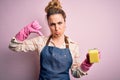Beautiful blonde cleaner woman doing housework wearing arpon and gloves using scourer with angry face, negative sign showing
