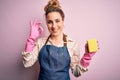 Beautiful blonde cleaner woman doing housework wearing arpon and gloves using scourer doing ok sign with fingers, excellent symbol