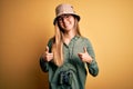 Beautiful blonde explorer woman with blue eyes wearing hat and glasses using binoculars success sign doing positive gesture with